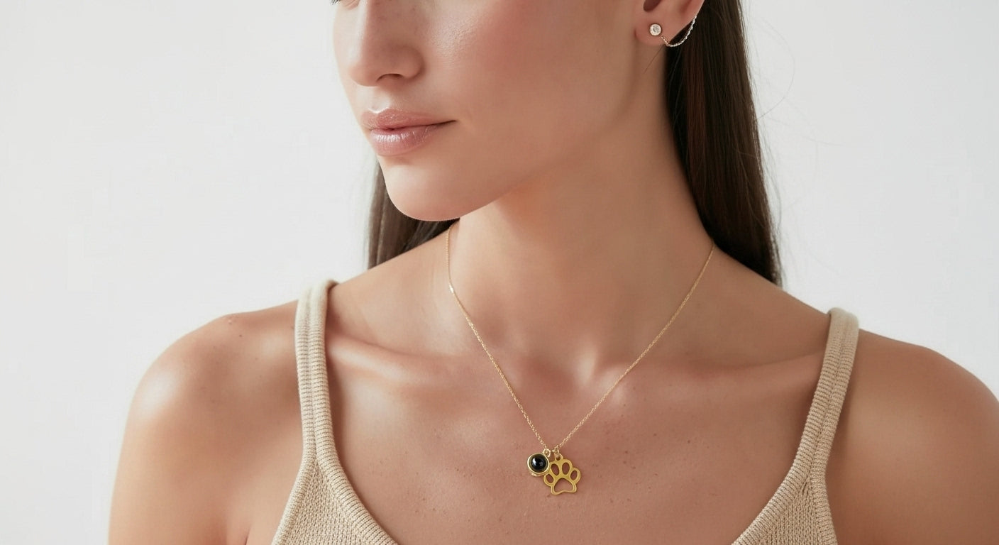 A close-up photograph of a woman wearing a delicate gold chain necklace from LumiPaw, featuring two pendants: a stylized gold paw print and a small circular projection lens for hidden pet photos. This custom necklace is a perfect personalized gift for pet lovers, specifically for dog and cat moms, and functions as meaningful pet loss or remembrance jewelry. Carry your pet's light always.